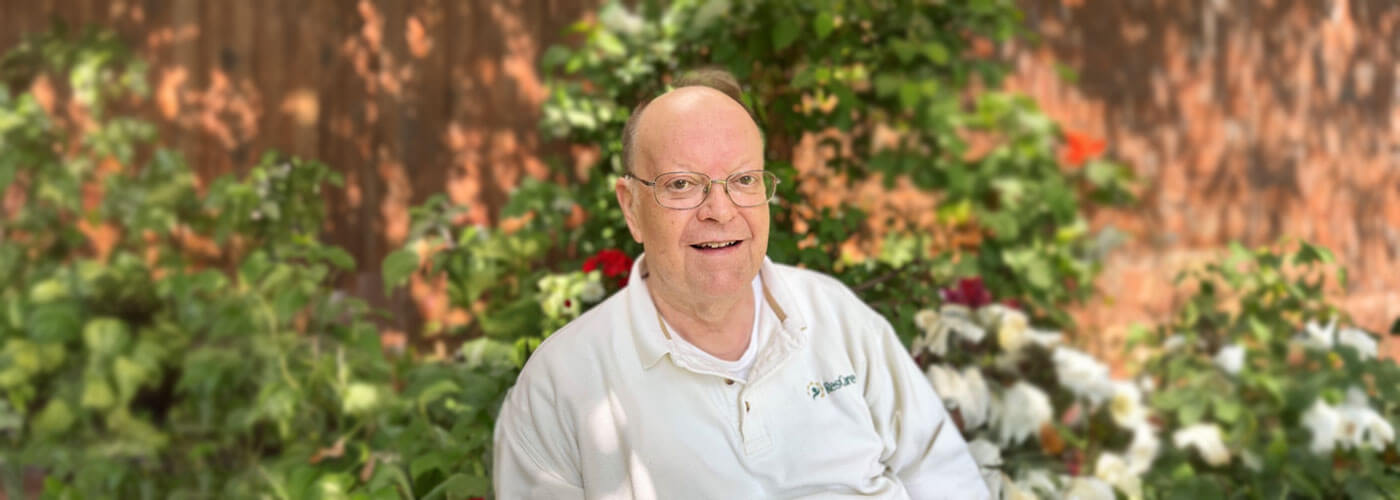 Senior man sitting in front of a brick wall and plants, smiling with glasses and a white collard shirt.