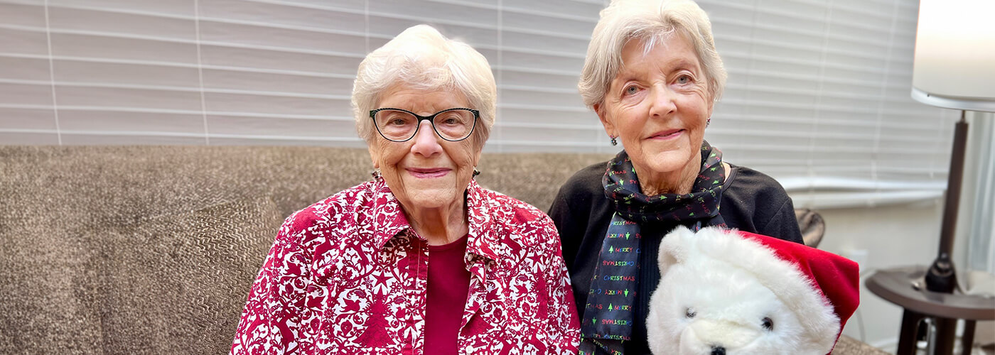 Two senior woman sitting on a tan couch smiling at the camera while holding a stuffed bear with a read holiday hat.