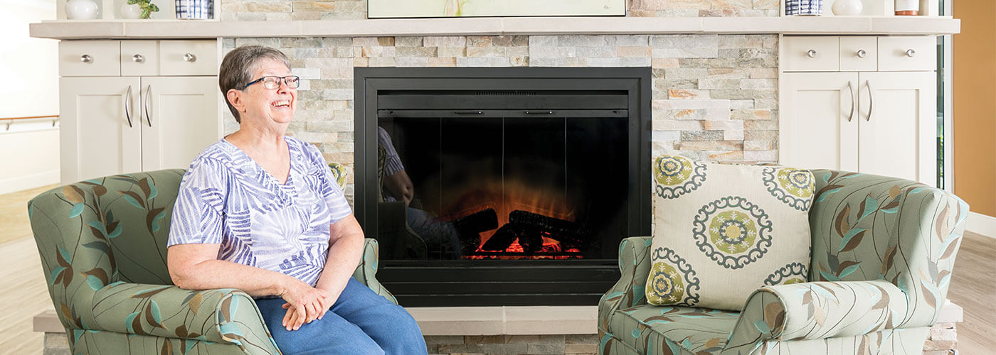 Westerwood resident, Remona, sitting in a chair laughing next to a fireplace.